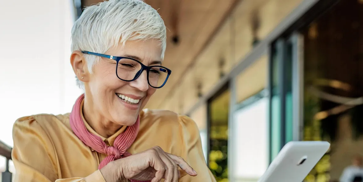 Mujer mayor con gafas sonriendo mientras usa una tableta digital en un espacio exterior moderno